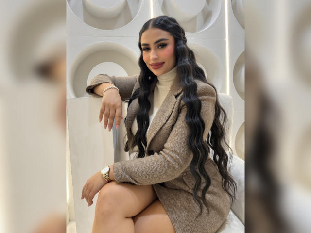 MiilaWx with long, wavy black hair, wearing a brown, woolen, button-up, knee-length coat, sitting, showing her ass, with a watch on her wrist, smiling, red lipstick.
