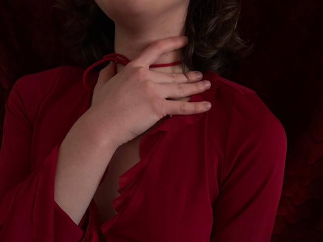 MiyaLia's hand grips her red shirt, fingers near her neck. Her shirt is open, showing a hint of her tits. Her brown hair is curled. Dark red background.