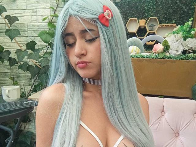 KatheLux with long, blue-green hair, a red flower clip, and a checkered choker, pouting with eyes closed, showing her big tits in a white strappy top.