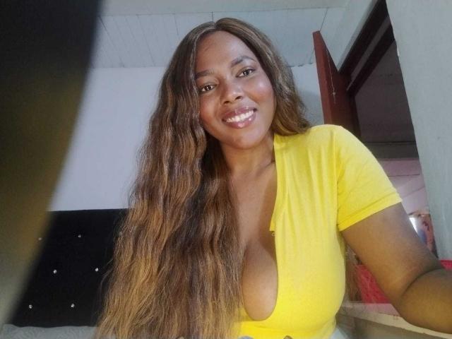 MacarenaBoobs has long, wavy brown hair, a yellow top with a deep cleavage, and is smiling at the camera, showing her shiny, white teeth. Her tits are noticeable.