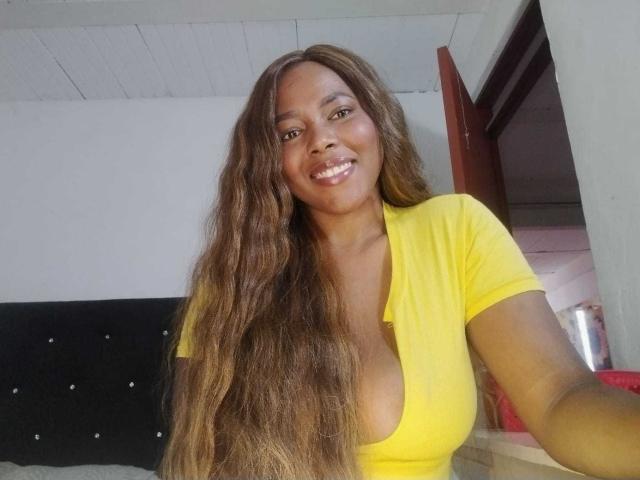 MacarenaBoobs with long wavy brown hair, yellow top, and big tits, smiling at the camera, standing in a room with a black headboard and open door.