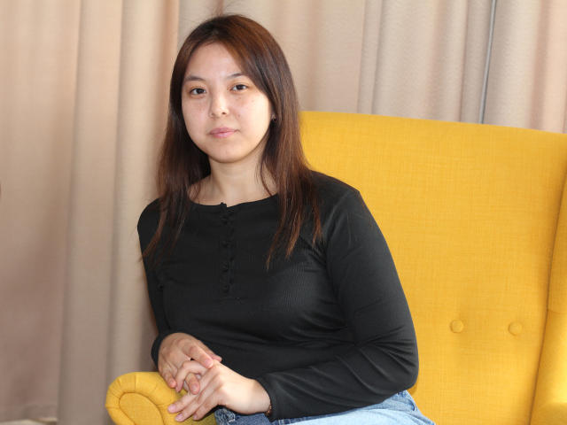 TeresaDouglas with medium tits, brown hair, black long-sleeve shirt, hands on yellow chair, beige curtain background, slight smile, casual pose, light skin, relaxed, sitting, looking at camera.