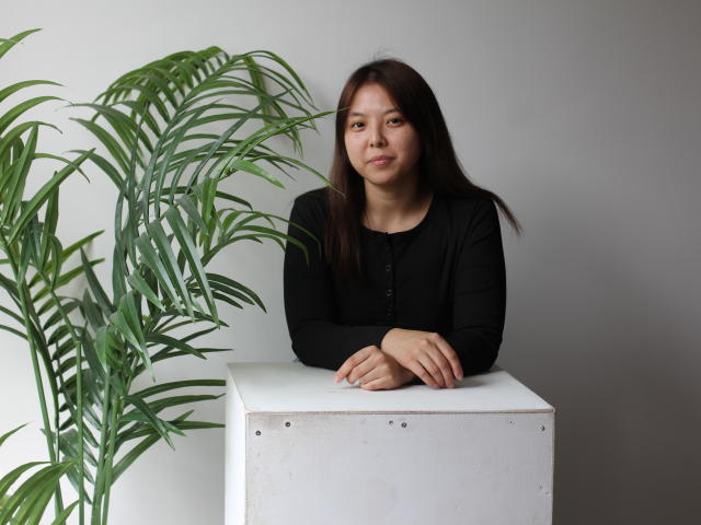 TeresaDouglas, with medium tits, long brown hair, and a black long-sleeve shirt, leans on a white cube, arms crossed, next to a green plant.