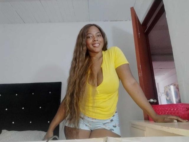 MacarenaBoobs with long brown wavy hair, yellow V-neck top, and blue denim shorts, smiling, leaning on bed, white walls, red door, black tufted headboard, and red bedspread.