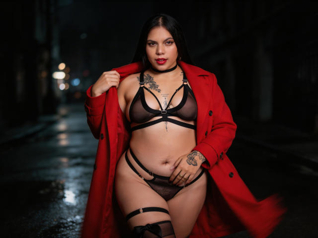 AuroraRosse, vestindo um casaco vermelho, lingerie preta transparente e tiras, em uma rua molhada à noite, exibindo seus seios, vulva e tatuagens, com um olhar confiante..