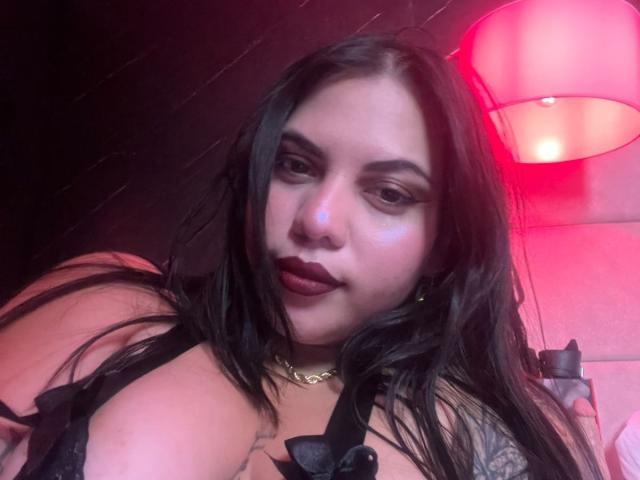 AuroraRosse met lang zwart haar, donkere lipstick en een gouden ketting, draagt zwarte lingerie, waarbij haar grote borsten zichtbaar zijn, in roze-rode verlichting, en kijkt naar de camera..