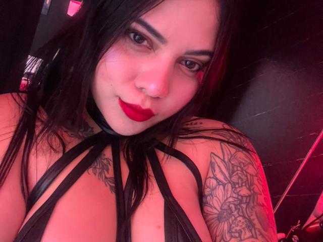 AuroraRosse, com cabelo escuro, batom vermelho, top preto com tiras, tatuagens no braço e seios grandes, olhando para a câmera com um leve sorriso. Luz vermelha ao fundo..