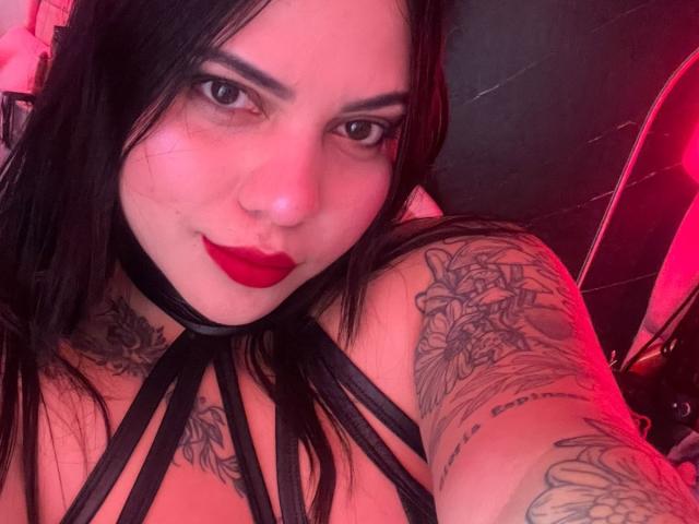 AuroraRosse, com cabelo preto, batom vermelho, tatuagens nos braços e no peito, vestindo uma blusa preta com alças finas, olhando para a câmera, iluminação vermelha, sorrindo, mostrando os seios..