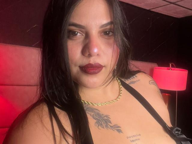 AuroraRosse con capelli scuri e umidi, rossetto rosso, canotta nera, catena d'oro, tatuaggi sul petto, espressione seria, lampada rossa sullo sfondo, che guarda nella fotocamera..