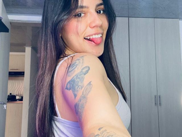 SophiaMarx with long brown hair, tongue out, white tank top, tattooed arm, smiling, showing off her tits and ass in a selfie, standing in a room with gray cabinets.
