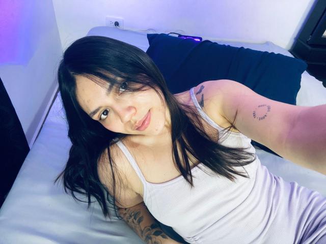 SophiaMarx, with long black hair, white tank top, tattoo on right arm, sitting on bed, smiling at camera, showing small tits, light skin, purple eyes, slight acne.