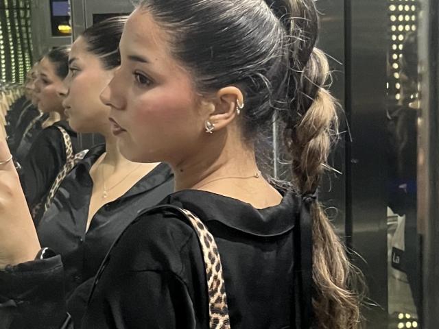 AmbarDunn with her brown ponytail, wearing a black shirt, looking in a mirror, showing off her small tits and blushed cheeks, with a leopard print bag.