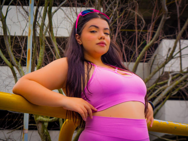 LaraJames with long black hair, pink sports bra showing her tits, pink high-waisted pants, pink headband, leaning on yellow bar, looking confident, tan skin, full lips.