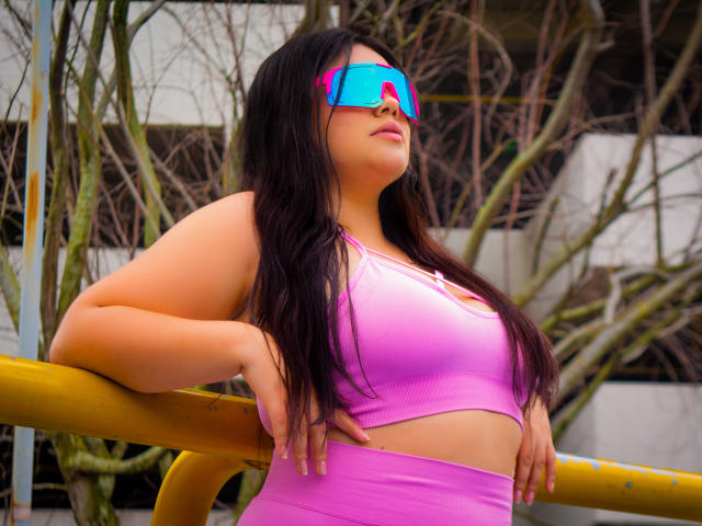 LaraJames with pink sports bra, matching pink bottoms, blue reflective sunglasses, dark wavy hair, hands on yellow bar, medium tits, confident pose, urban background with leafy branches.