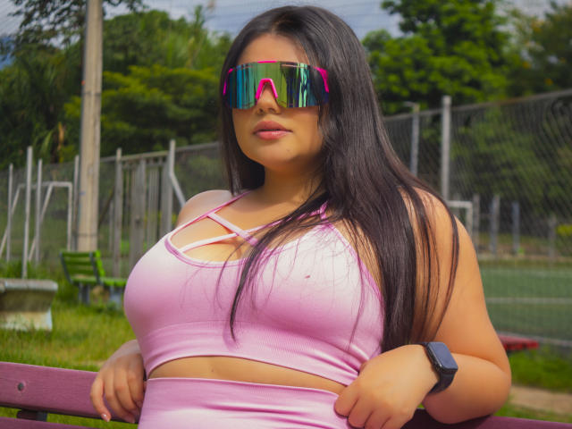 LaraJames with long black hair, pink sports bra, and matching pink shorts, wearing reflective pink and green sunglasses, standing with hands on a purple bench, wristwatch on.