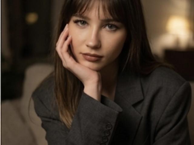 Adelisha with brown hair, bangs, and grey blazer, resting her hand on her face, looking slightly sad or contemplative. Her brown eyes are focused. She has a small nose.
