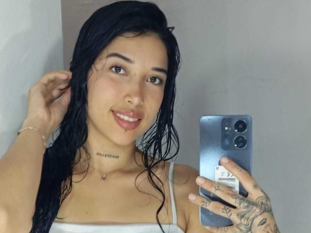 KrystalEvans with wet black hair, white tank top, nose ring, tattooed hand, and a phone selfie, smiling, showing pink lips, and a small tattoo on her neck.