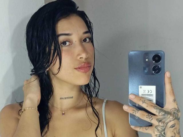 KrystalEvans with wet black hair, holding a phone, showing tattoos on her hand, wearing a white tank top, and a nose ring, with a slight pout.