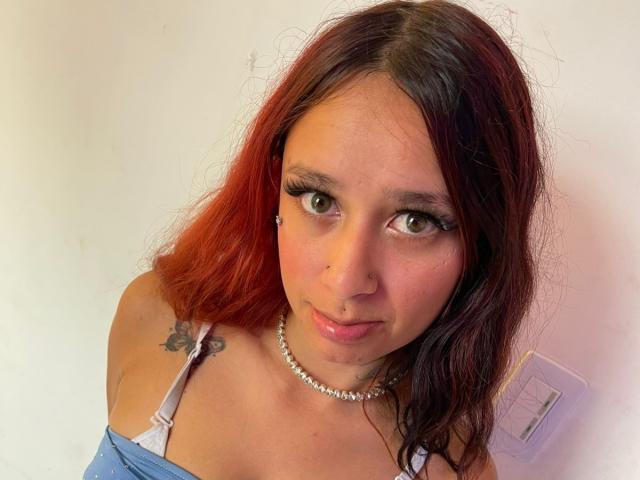 RoxanaSanchezz has red and brown wavy hair, green eyes, and a nose piercing. She's wearing a pearl necklace and a blue-off-shoulder top with visible tits and tattoos.