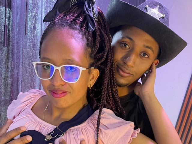 LilithAndAsmodeo with braided hair in a black bandana, white reflective glasses, pink dress, and red lipstick, stands next to LilithAndAsmodeo in a black shirt and cowboy hat.