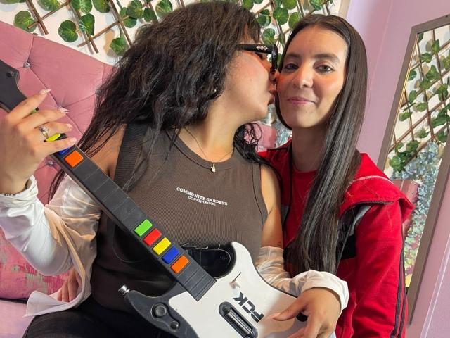 ValkiriaAndDione with long black hair kissing ValkiriaAndDione, holding a white electric guitar with colorful buttons. ValkiriaAndDione in brown sleeveless top, ValkiriaAndDione in red jacket.