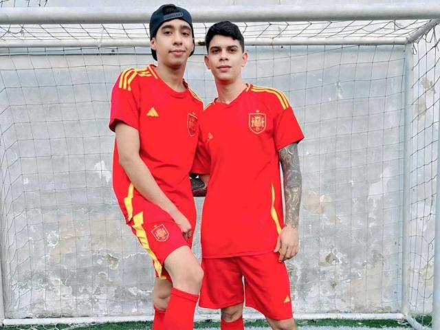 Two TeamArs in red soccer jerseys with yellow stripes, standing in front of a net. One has a black cap, the other has tattoos on his right arm.