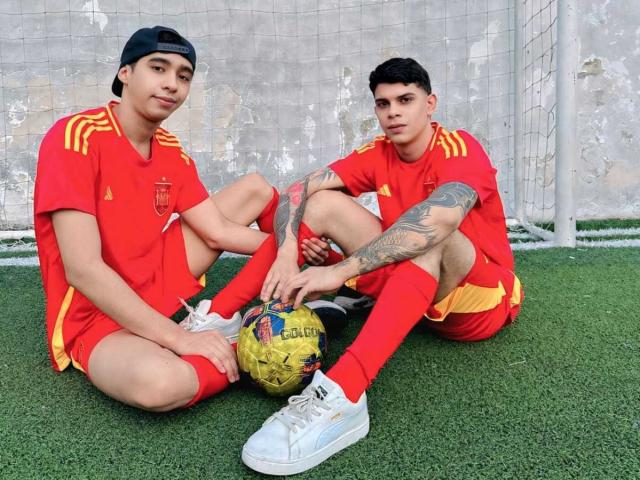 Two TeamAr dudes in red soccer jerseys and shorts, sitting on green turf, one with tattoos, both wearing white sneakers, black caps, and holding a yellow soccer ball.
