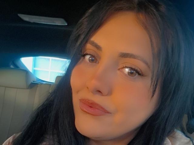 AlesiaLux has brown eyes, long black hair, and full pink lips. She's giving a slight pout with a subtle smile. Her makeup includes thick eyelashes and defined eyebrows.