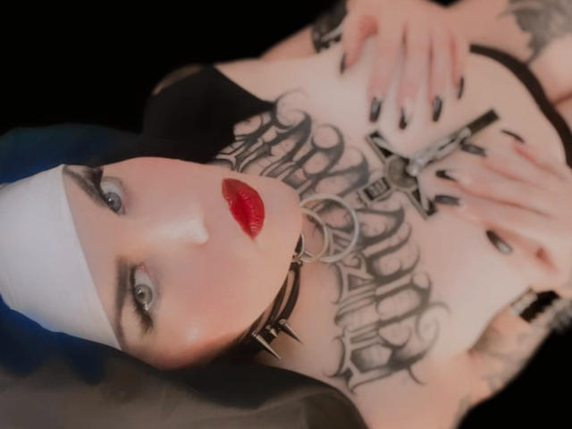 FetishHottieForU with red lipstick, blue hair, white headband, black choker, tattoos, and black nails, lying down, hands on chest, showing bare tits, black background.