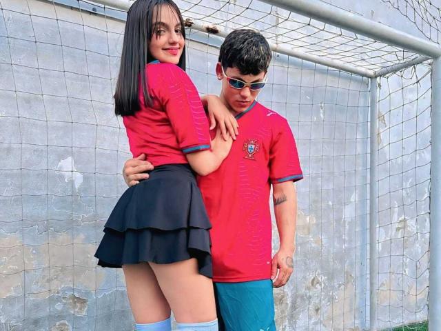 EndlessTwo with black hair and red soccer jersey, black skirt, and knee socks, stands with her ass to the camera, arms around a guy in a red soccer jersey.