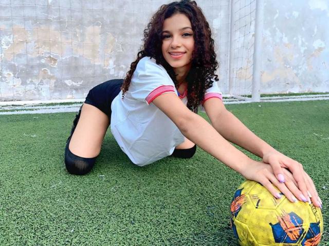 BellainLove, with curly brown hair, kneels on green turf, stretching over a yellow soccer ball, wearing a white shirt with pink trim, black shorts, and black knee socks.