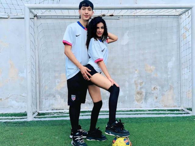 BellainLove with black hair, white soccer shirt, black shorts, and knee socks, stands with her hand on her ass, next to a boy in a white soccer shirt, on green grass.