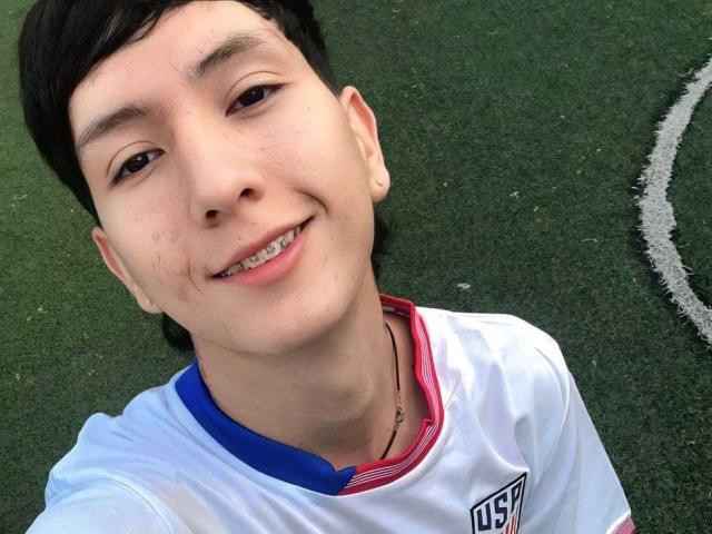 BellainLove, a young dude with short black hair, slight acne, and a cute smile, wearing a white soccer jersey with blue and red trim, on a green turf.