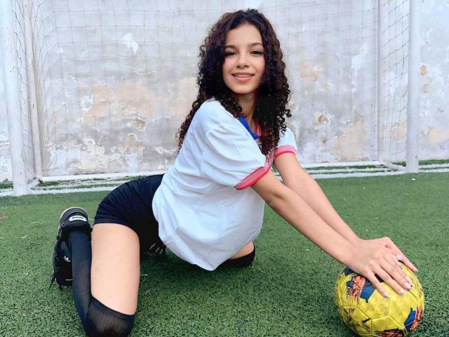 BellainLove with curly brown hair, white soccer shirt, pink trim, black shorts, black knee socks, kneeling on green grass, holding yellow soccer ball, smiling, hands on ball.