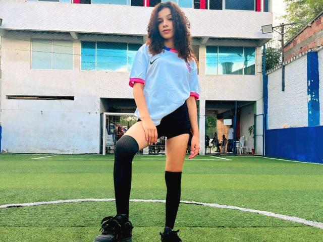 BellainLove has curly brown hair, wears a light blue soccer shirt with pink sleeves, black shorts, knee socks, and black shoes, standing on a green soccer field.