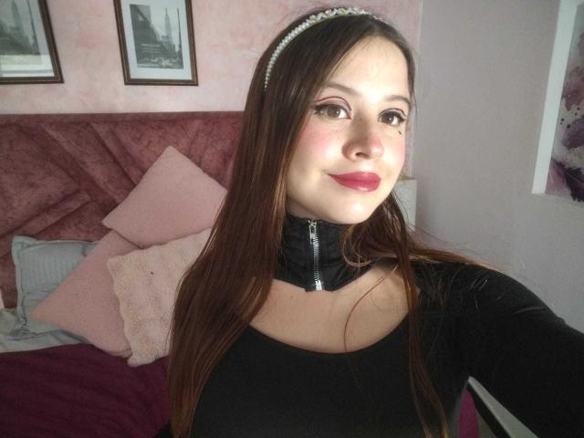 MelaniBecker with long brown hair, black zip-up top, pink lipstick, and headband, smiling, showing her tits, in a pink and purple bedroom.