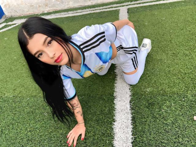 TheXPleasure, mit langem, schwarzem Haar, rotem Lippenstift und Tätowierungen, kniet auf einem grünen Fußballfeld, trägt einen weißen und blauen Fußballanzug, zeigt ihr Gesäß und blickt nach oben..