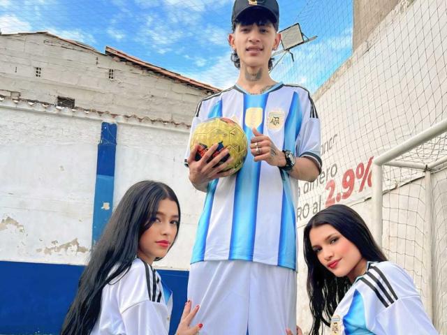 TheXPleasure, een lange kerel met een baseballpet, die een gele voetbal vasthoudt, gekleed in een blauw en wit Argentijns voetbalshirt, omringd door twee aantrekkelijke, donkerharige Latina's in vergelijkbare shirts..