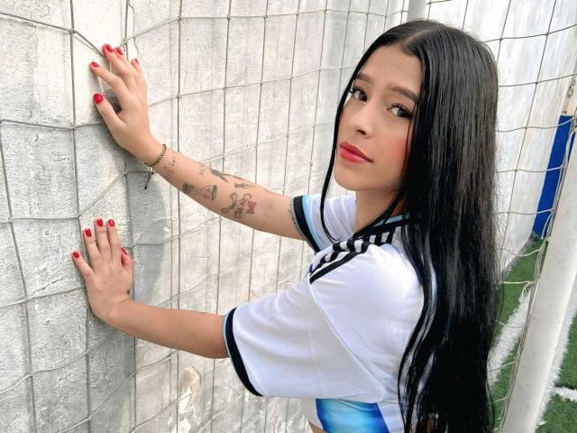 TheXPleasure has long black hair, red lipstick, and tattoos on her right arm. She's wearing a white soccer jersey, red nails, and is leaning against a silver wall.