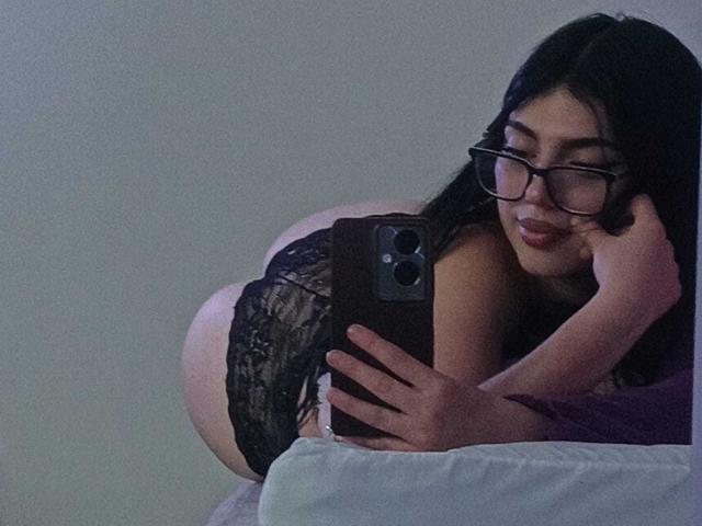IsabellaaRossi in black lace panties, on knees, butt up, taking a selfie with glasses, phone in hand, smiling, on a bed with purple sheets, in dim light.