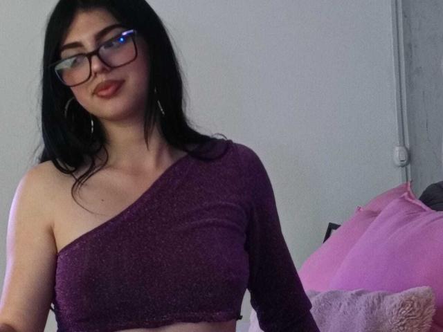 IsabellaaRossi with black hair, glasses, and hoop earrings, wearing a purple one-shoulder crop top, smiling. Boobs visible under the top, pink pillows in the background.