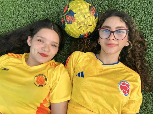 Dos niñas con cabello castaño rizado, vestidas con camisetas de fútbol amarillas, acostadas sobre el césped, sonriendo a un balón de fútbol amarillo con patrones naranja y azul. Ambas llevan lápiz labial rojo..