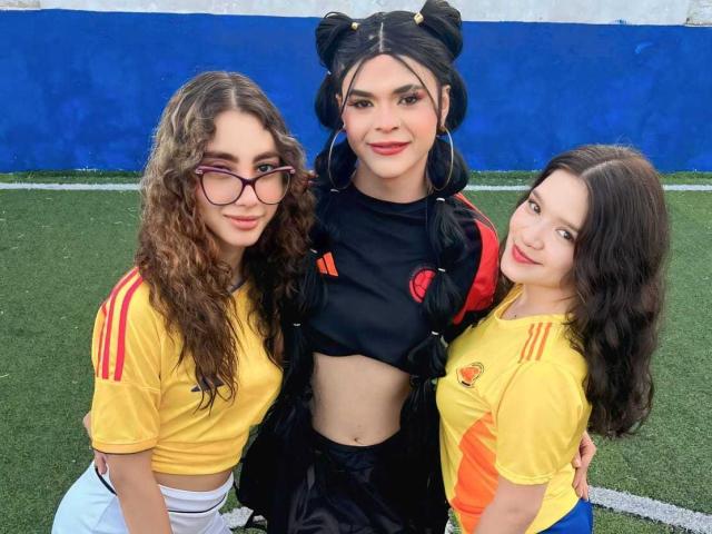 Tres mujeres con cabello oscuro, rizado y lacio, "PowerofThree" vestidas con una camiseta negra con mangas rojas y negras, usando camisetas amarillas con rayas rojas, de pie sobre césped verde..