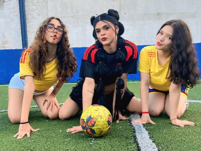 Three PowerofThrees on fake grass, knees on soccer field, yellow shirts, black and red, black pigtails, curly brown hair, glasses, shorts, soccer ball, serious and playful expressions.