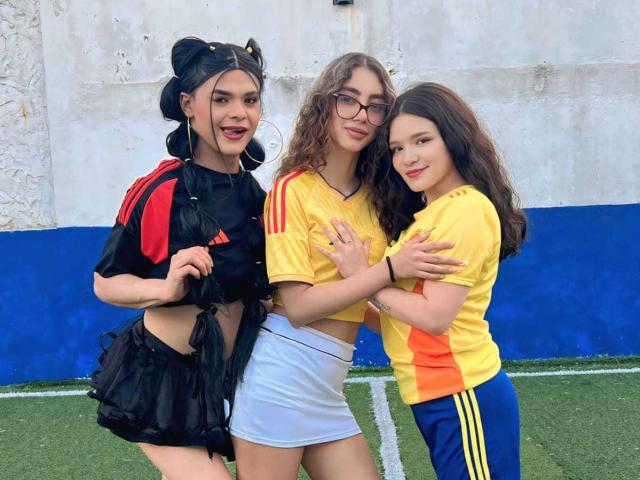 Tres personas de PowerofThrees están de pie sobre el césped. Una lleva un top deportivo negro y rojo, una falda negra y moños de pelo negros. Las otras dos llevan camisetas deportivas amarillas y naranjas, gafas y faldas blancas..