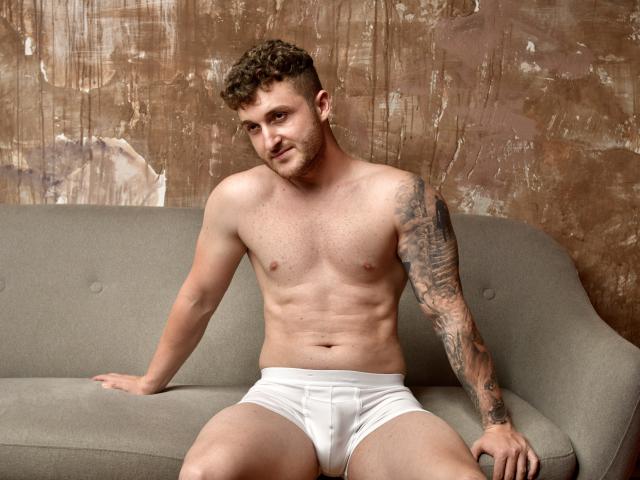 EthannI with curly brown hair, tattoos on his right arm, and white boxer briefs, sitting on a gray couch, showing a muscular, hairy chest and a slight smile.