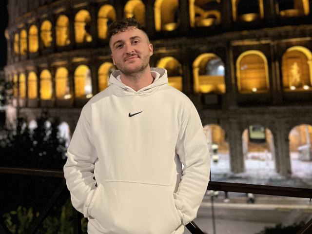 EthannI, con cabello castaño rizado, barba clara, vistiendo una sudadera blanca Nike, con las manos en los bolsillos, de pie frente a un edificio iluminado por la noche..