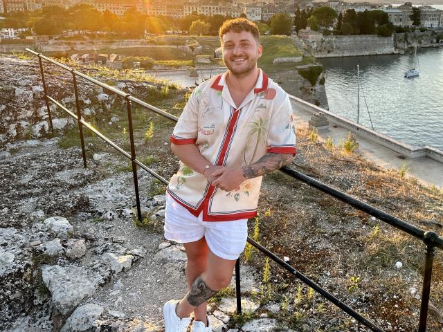 EthannI has a cute, smiling face, tattooed arms, and a hairy chest. He's wearing a floral shirt, white shorts, and white sneakers, standing by a metal railing.