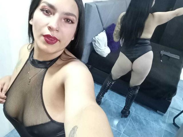 KiaraThomsomp portant un haut en mesh noir à encolure plongeante, qui met en valeur ses seins et ses fesses, vêtue d'un string noir et de bottes hautes, se prenant en selfie, reflet dans un miroir, rouge à lèvres rouge, piercing au nez..