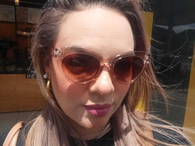 AvaMia with tan skin, pink lipstick, and purple eyeshadow, wearing large brown sunglasses and gold hoop earrings, has long brown hair and a black top.